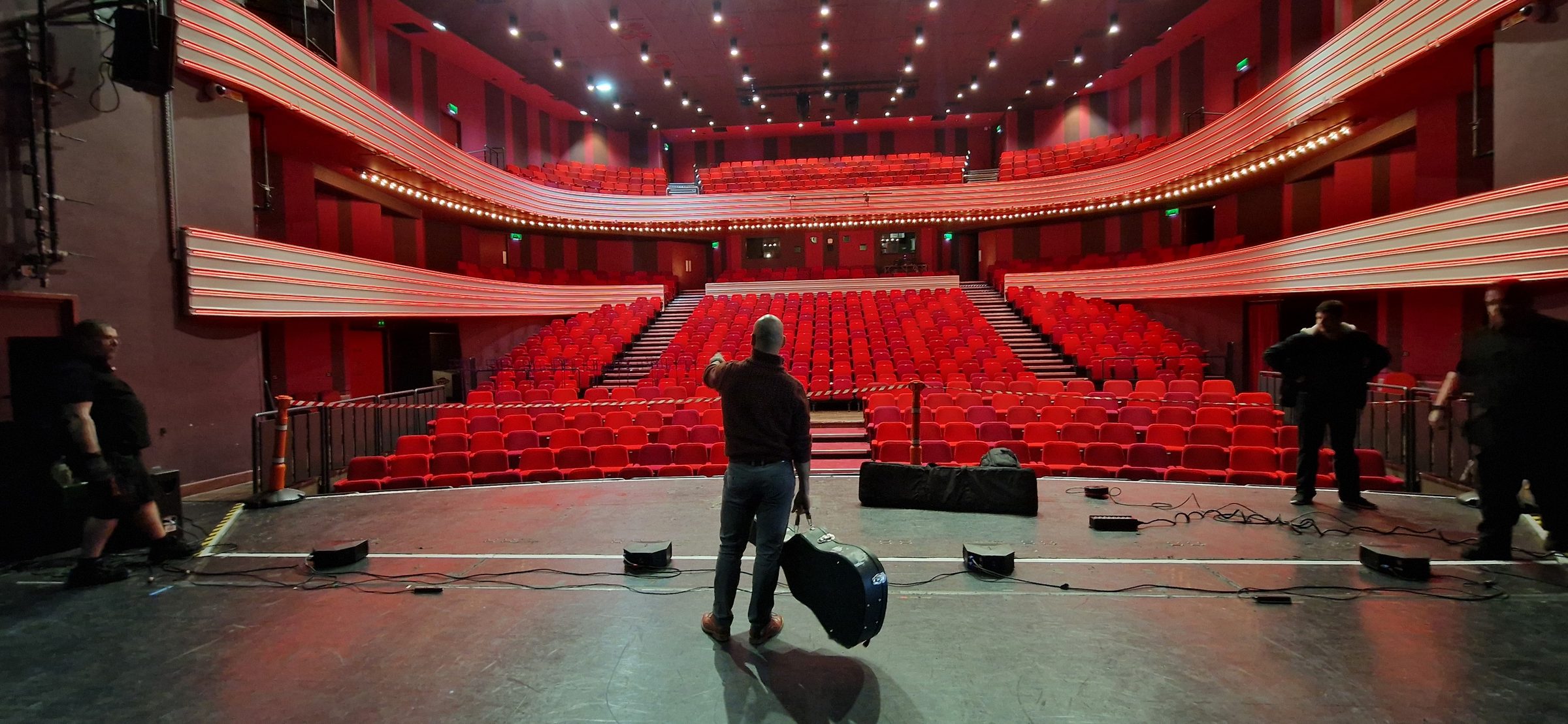 Barry de Jager standing alone on a theatre stage, guitar case in hand, facing rows of empty red seats