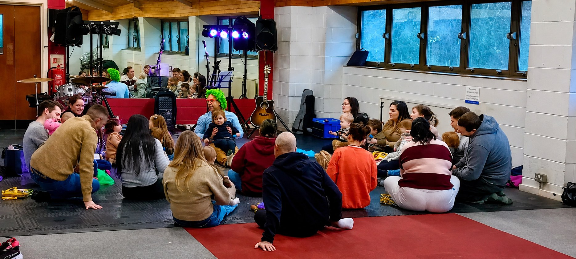 Parents, carers and babies gathered in a communal circle on the floor during a live Baby Concert — Barry de Jager sits among them with stage lighting, guitars and drums behind