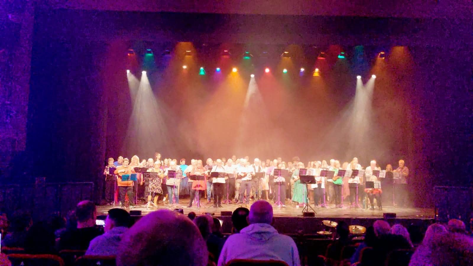 A large ensemble of ukulele players performing together on a theatre stage under warm lights