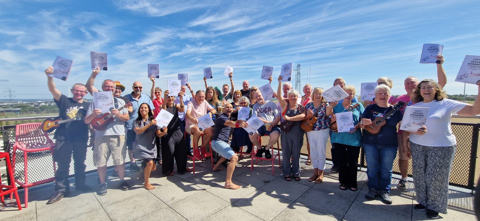 A large outdoor group holding copies of Barry de Jager’s Ukulele Songbook up in the air