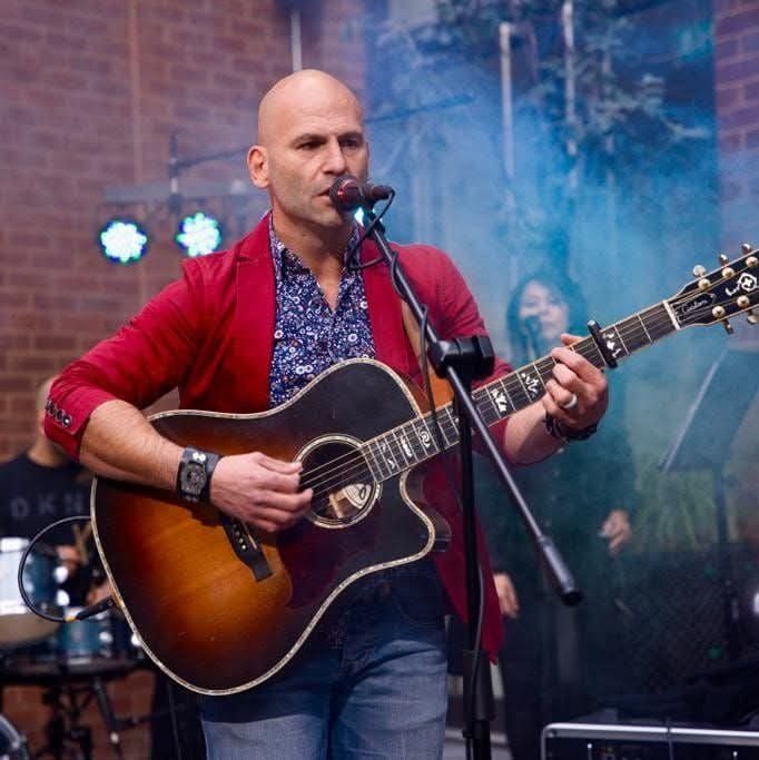 Barry de Jager performing live on stage with an acoustic guitar under coloured lights